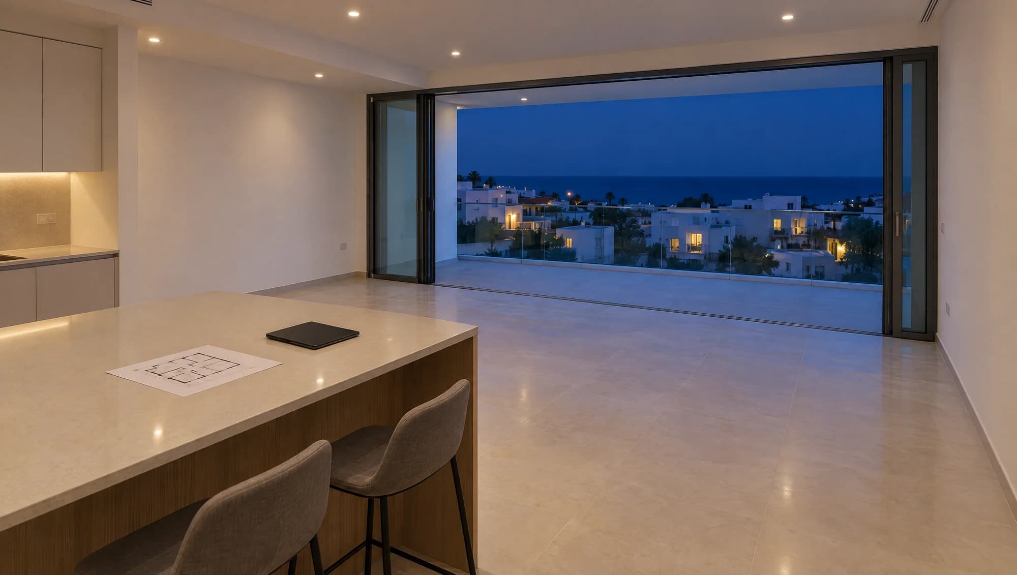 An empty new flat at dusk: a kitchen island with a floor plan, sliding doors open onto a terrace with a sea view